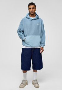 Lichtblauw hoodie met een buidelzak en wit logo, gecombineerd met donkerdenim shorts en beige sneakers. Witte sokken zichtbaar.