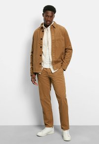 Mann in beige Kord-Jacke und -Hose über einem weißen Hemd mit weißen Sneakern, der vor einem schlichten hellen Hintergrund steht.
