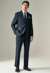 Marineblauer karierter Anzug mit abgesetztem Revers, passenden Hosen und einem weißen Hemd. Schwarze Schnürschuhe und ein gemusterter Einstecktuch.
