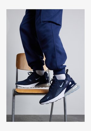 Personne portant des baskets Nike Air Max 270 bleu marine et un pantalon de survêtement bleu marine assorti, debout sur une chaise en bois avec des pieds en métal.