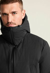 Schwarze isolierte Jacke mit hohem Kragen und Kapuze, ausgestattet mit einem durchgehenden Reißverschluss und minimaler Fronttasche. Glatte Textur, modernes Design.