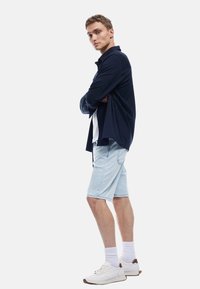 Camisa de botones azul marino, superpuesta sobre una camiseta blanca, combinada con pantalones cortos de mezclilla azul claro y zapatillas blancas con acentos en marrón. Medias blancas visibles.