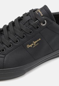 Sapatilhas pretas com amarração, com um upper em couro texturizado, sola em borracha e a marcação em dourado "Pepe Jeans London". Apresenta ilhós e atacadores pretos.