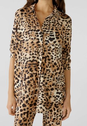 Frau trägt passend gemustertes Leoparden-Longsleeve-Button-up-Hemd und Hose mit hochgekrempelten Ärmeln vor schlichtem Hintergrund.