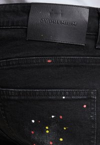 Czarne dżinsy z denimu z skórzaną metką oznaczoną "ZY PREMIUM". Mają wielokolorowe plamy farby na tylnej kieszeni. Klasyczny pięciokieszeniowy krój.