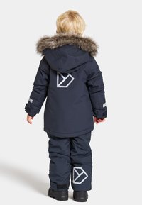 Manteau d'hiver marine avec capuche en fourrure, accents réfléchissants et détail de logo dans le dos, associé à un pantalon marine assorti et des bottes noires.