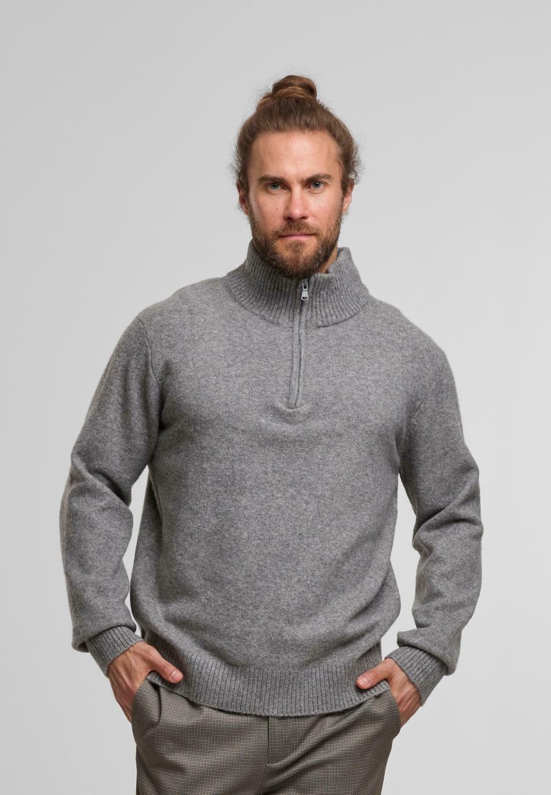 Light grey zip-neck sweater made of soft, textured fabric. Features ribbed collar and cuffs, and a relaxed fit. Paired with checkered trousers.