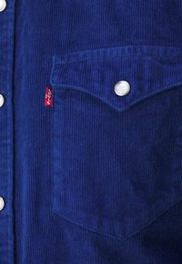 Blå kordflodtröja med knappdesign, silver färgade tryckknappar och en bröstficka med en röd Levi's etikett. Texturerad tyg synlig.