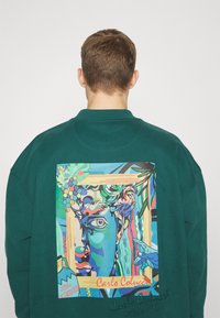 Groene sweatshirt met een kleurrijk kunstwerk dat een gezicht met druiven en handen afbeeldt, ingelijst op de achterkant. Handtekening in de hoek.