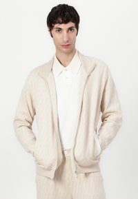 Veste beige zippée avec un motif en losanges texturé, dotée d'un col et de poignets côtelés. Portée sur une chemise blanche à col. Pantalon assorti.