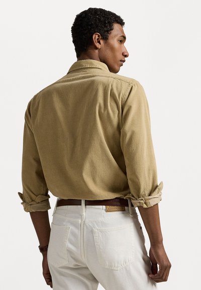 Kaki corduroy skjorte med oppbrettede ermer, med en klassisk krage og tekstur, kombinert med hvite jeans og et brunt lærbelte.