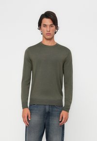 Olive zelena, lahka pletena jopica z dolgimi rokavi in krožnim izrezom, v kombinaciji z modrimi denim hlačami z rahlim bledenjem.