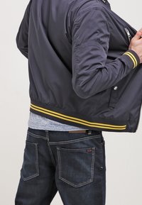 Giacca bomber di un blu navy scuro con strisce gialle all'orlo, tasche con zip e polsini a costine, indossata sopra un top azzurro chiaro e jeans di denim.