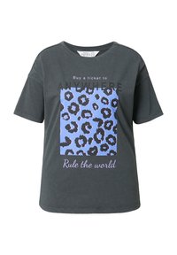 Camiseta de algodón gris con mangas cortas, que presenta un cuadrado de estampado de leopardo azul y texto negro: “Compra un billete a CUALQUIER LUGAR” y “Domina el mundo.”