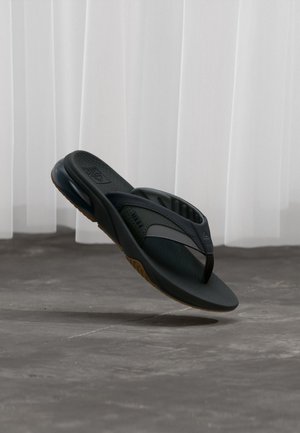 Must papuga sandaal polsterdatud taldsete ja tekstuurse sisetallaga, hõljumas halli põranda kohal valgete kardinate ees.