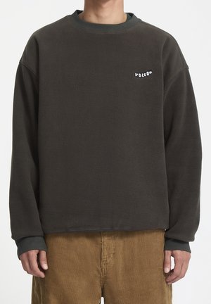 Osoba ubrana w ciemnoszarą bluzę Volcom z polaru i brązowe spodnie sztruksowe, stojąca z rękami swobodnie opuszczonymi wzdłuż ciała.