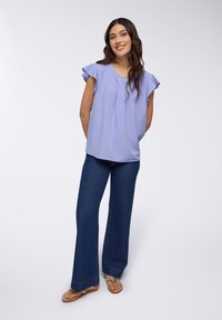 Blusa viola chiaro con maniche a cappuccio, abbinata a jeans svasati in denim scuro. Le scarpe consistono in infradito color sabbia con cinturini decorati.