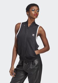 Gilet sans manches noir avec des accents blancs, fermeture zippée, col haut et deux poches latérales, fabriqué en tissu athlétique lisse.