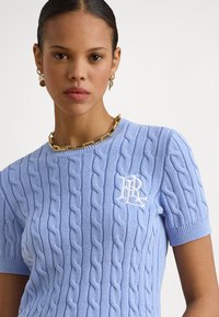 Svetlo modra pletena majica s kabelskim vzorcem, kratkimi rokavi in belim logotipom na levi strani prsi. Zlata verižica dopolnjuje dizajn.