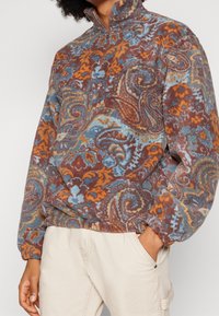 Pullover in pile con mezza zip, caratterizzato da un motivo paisley in tonalità di marrone, blu e arancio. Morbida consistenza con polsini a coste.