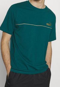 Turkusowa bawełniana koszulka z złotą poziomą paskiem, z logo Pumy na lewej piersi. Krótkie rękawy i okrągły dekolt.