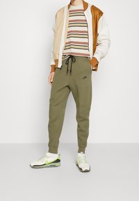 Sportovní oblečení zahrnující béžovo-hnědou bomber bundu, proužkované béžovo-vínové tričko, olivově zelené tepláky a bílé tenisky Nike.