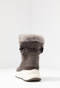 Stivale alla caviglia in suede marrone con bordo in pelliccia chiara, cinturino regolabile nero e suola spessa bianca su sfondo bianco.
