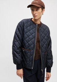 Jūras spēku šūta bomber jaka ar brūnu apdari un kabatām, ar priekšējo rāvējslēdzēju un ribotu aproču. Valkāta pār brūnu džemperi.