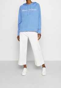 Felpa blu con cappuccio e logo frontale, abbinata a pantaloni cropped larghi bianchi e sneakers bianche; presenta un tessuto morbido e una vestibilità rilassata.