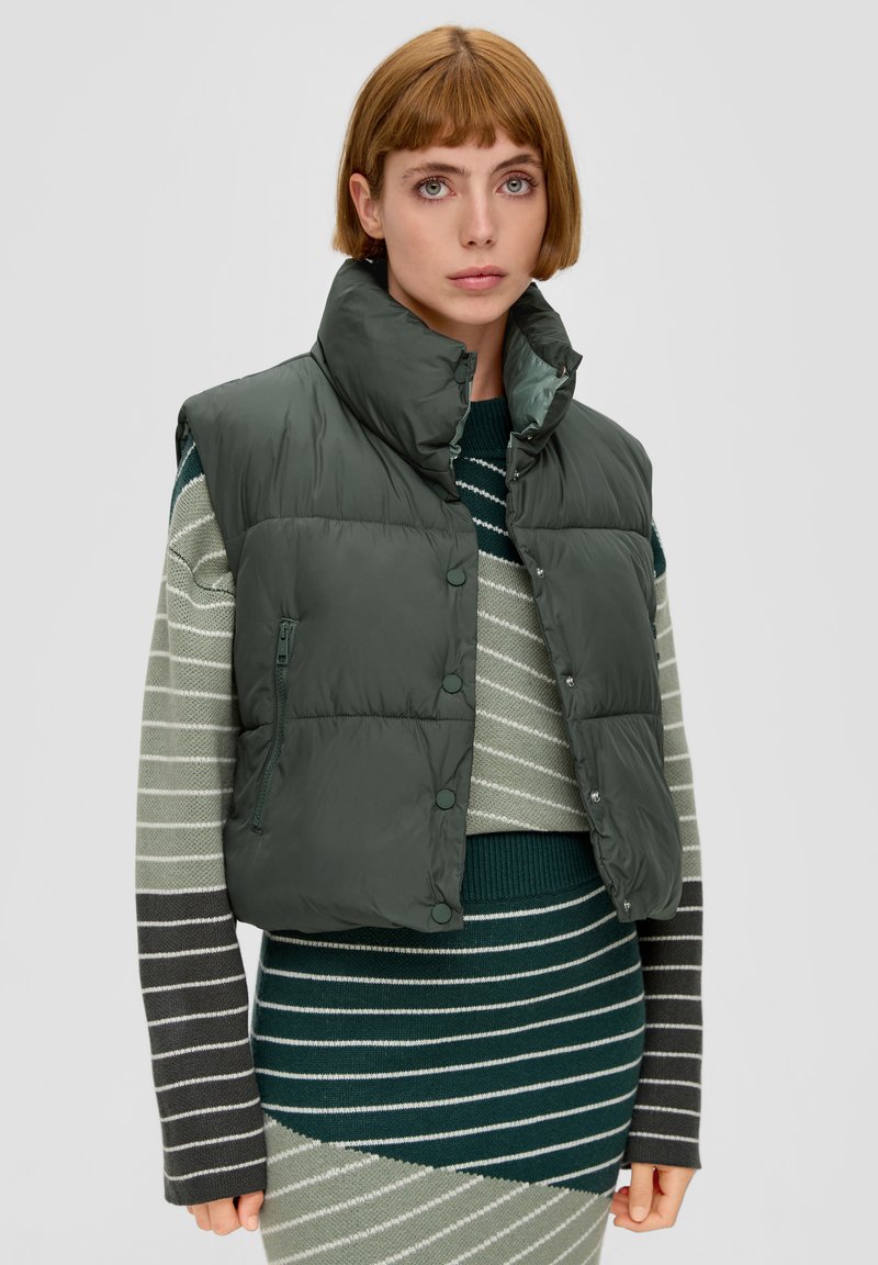 Dunkelgrüne Steppweste mit hohem Kragen, Druckknöpfen und einer seitlichen Reißverschlusstasche. Getragen über einem gestreiften Strickpullover.