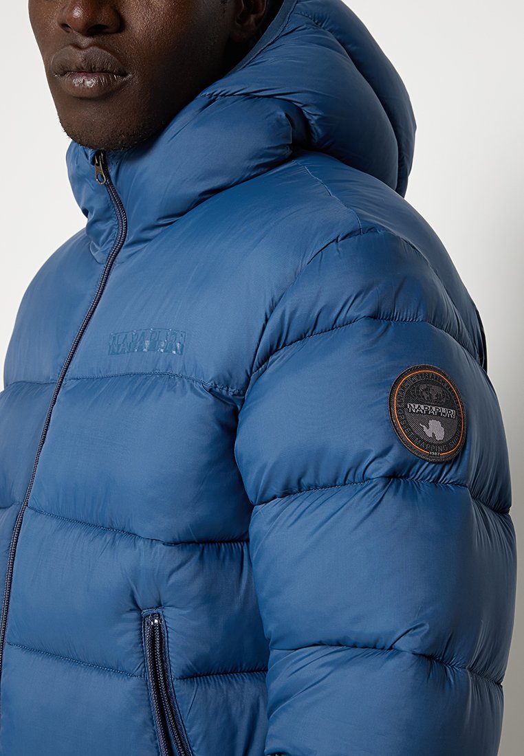 Chaqueta acolchada azul con capucha, textura acolchada, cierre de cremallera y bolsillos laterales. Presenta un logotipo de marca circular en la manga izquierda.