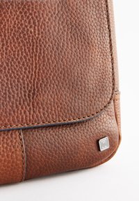 Geantă maro din piele cu suprafață texturată, având o margine cusută și un logo argintiu metalic în colțul din față.