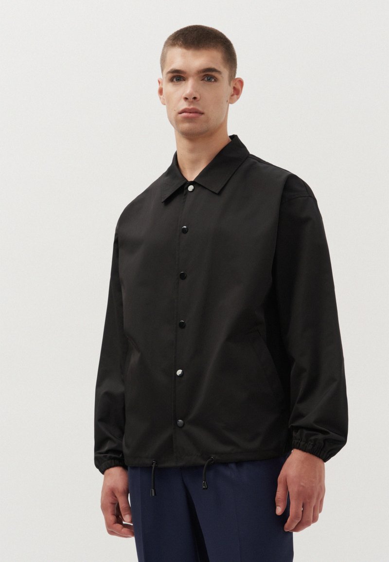 Veste noire légère avec un col, des boutons pression, des poignets élastiques et un ourlet ajustable. Texture lisse, design minimaliste.