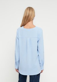 Φωτεινό γαλάζιο μπλούζα με μακριά μανίκια από υφή υφάσματος, στρογγυλή λαιμόκοψη και μανσέτες με κουμπιά. Διαθέτει πλέξη στην πλάτη για επιπλέον λεπτομέρεια.