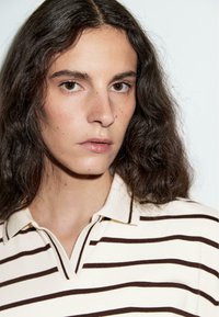 Crème polo shirt met donkerbruine horizontale strepen, voorzien van een platte kraag en korte mouwen, gemaakt van een zachte stof.
