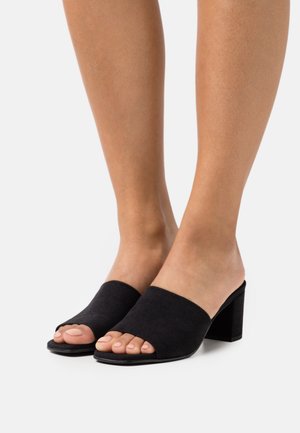 Mules ouvertes en daim noir avec une large bride sur le dessus du pied et un talon épais, présentant une texture lisse et un design minimaliste.