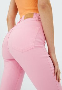 Rosa Jeans aus Denim mit tailliertem Design, featuring doppelte Gesäßtaschen und einem braunen Leder-Logopatch an der Taille. Glatte Textur.