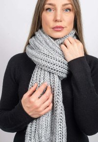 Grå stickad scarf gjord av tjockt garn, med ribbad textur och en lång, hängande design. En person håller sjalen.