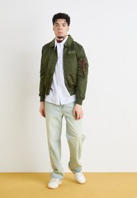 Oljčno zelena bomber jakna z ovratnikom, žepki z zadrgo in etiketo; kombinirana s svetlo modrimi kavbojkami z ravnimi hlačnicami in belimi supergami.