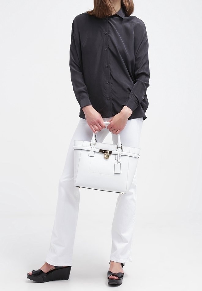 Chemise noire à manches longues, pantalon blanc, sandales compensées noires, et un sac à main blanc structuré avec un accent doré et une étiquette amovible.