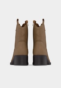 Ζευγάρι ταμπά μποτάκια από σουέντ με μαύρα στιβαρά τακούνια και χρυσά φερμουάρ στο πίσω μέρος, εμφανιζόμενα από πίσω σε λευκό φόντο.