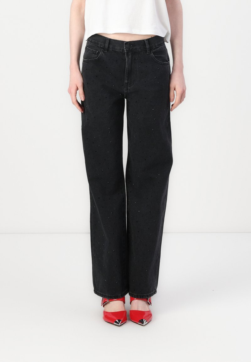 Black denim jeans with a loose fit, featuring small sparkling details throughout. Tapered leg design and five-pocket style, worn with red shoes.
