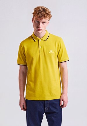 Junger Mann mit lockigem Haar, der ein gelbes Poloshirt und dunkelblaue Jeans trägt, steht vor einem schlichten hellen Hintergrund.