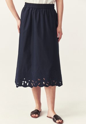 Jupe midi bleu marine avec ceinture élastique et broderie florale ajourée le long de l’ourlet, portée avec des sandales en cuir tressé marron foncé.