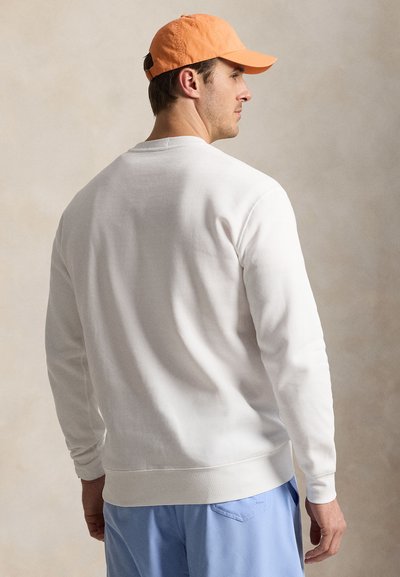 Oldalra néző férfi, narancssárga sapkát, fehér hosszú ujjú inget és világoskék nadrágot visel, semleges háttér előtt.