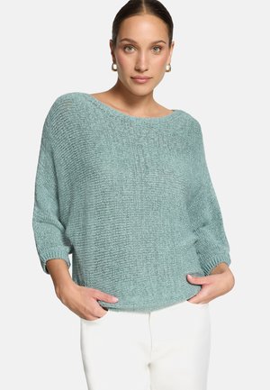 Femme portant un pull léger bleu clair à maille lâche avec des manches trois-quarts et un pantalon blanc, debout les mains dans les poches.