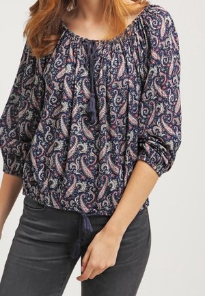 Femme portant un chemisier bleu marine à motif paisley avec un col froncé et des pompons, associé à un pantalon gris foncé ajusté.
