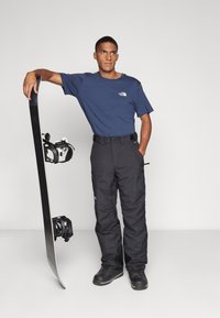 Snieglentėjas, vilkintis mėlyną „The North Face“ marškinėlį ir juodus sniego kelnes, stovintis šalia snieglenčių su tvirtinimais, prieš baltą foną.