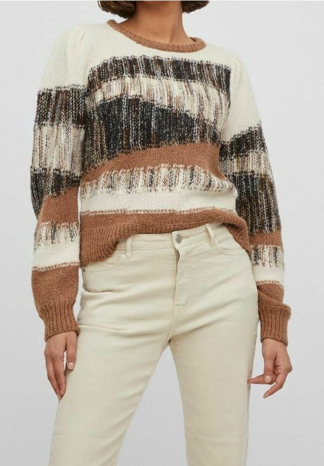 Strickpullover in creme, braunen und schwarzen Streifen. Verfügt über einen strukturierten Stoff und Rippbündchen. Kombiniert mit weiß-beigen eng anliegenden Hosen.