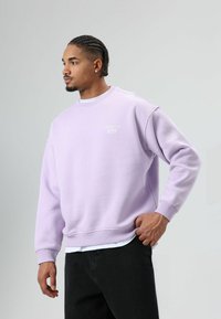 Sweat-shirt violet clair à coupe décontractée, avec poignets et ourlet côtelés. Présente un logo blanc sur la poitrine, associé à un pantalon noir.